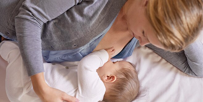 Eine Mutter liegt auf ihrer linken Seite auf einem weißen Bett und wiegt ihr Baby, das sie im Liegen stillt.