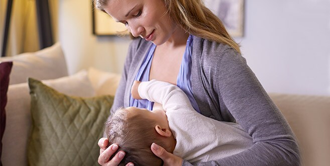 Eine Mutter sitzt aufrecht auf einem cremefarbenen Sofa und blickt auf ihr Baby hinunter, das sie mithilfe des Unterarmgriffs stillt.