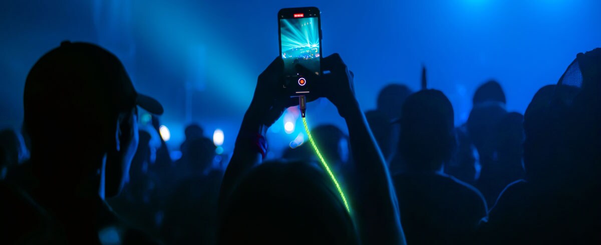 Eine Person hält ein Handy, das mit einem leuchtenden Kabel verbunden ist, vor einer großen Menschenmenge und fängt den Moment während einer Veranstaltung ein.