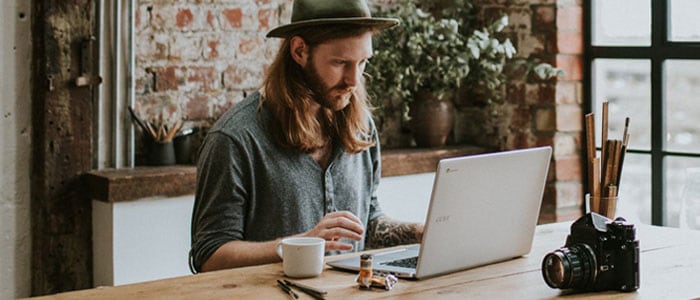 Ein Mann mit einem Hipster-Bart und langen Haaren sitzt vor einem Laptop.
