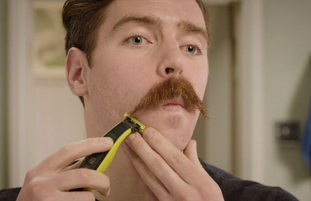 Gesicht eines Mannes mit einem Schnurrbart, der Barthaare auf der Wange mit einem elektrischen Rasierer trimmt.