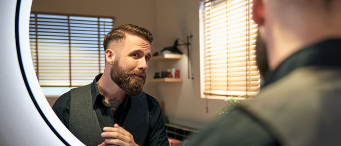 Ein Mann mit Bart und kurzem Haarschnitt lächelt sich im Spiegel an, nachdem er sich zu Hause die Haare geschnitten hat.